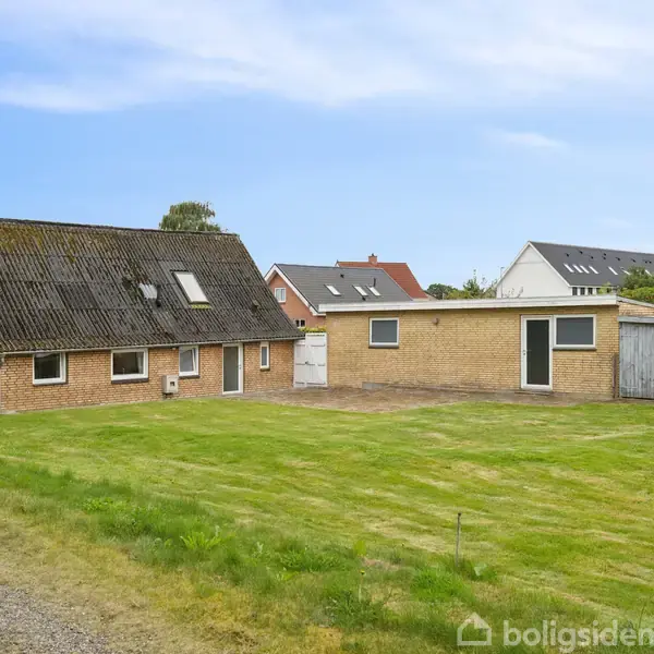 En brun murstensbygning med skråtag og flere vinduer står stille på en velplejet græsplæne. I baggrunden ses flere huse i et roligt villakvarter.