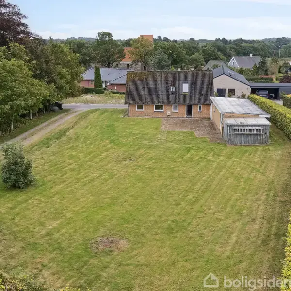 Et gult murstenshus står på en stor græsplæne. Ved siden af huset er en tilbygning med tag. Omkringliggende hække og træer omkranser ejendommen i et villakvarter.