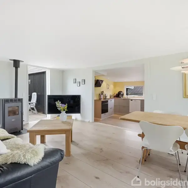 Sofa med puder og tæppe står ved brændeovn. Træbord med vase foran. Spisebord med stole ved siden af. Køkken med grå skabe i baggrunden. Lys og åben stue.
