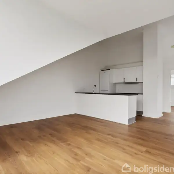 Et tomt rum med trægulv og hvidmalede vægge. Et moderne køkken med hvide skabe og mørk bordplade er indbygget. Rummet har et skrånende loft, hvilket skaber en rummelig fornemmelse.