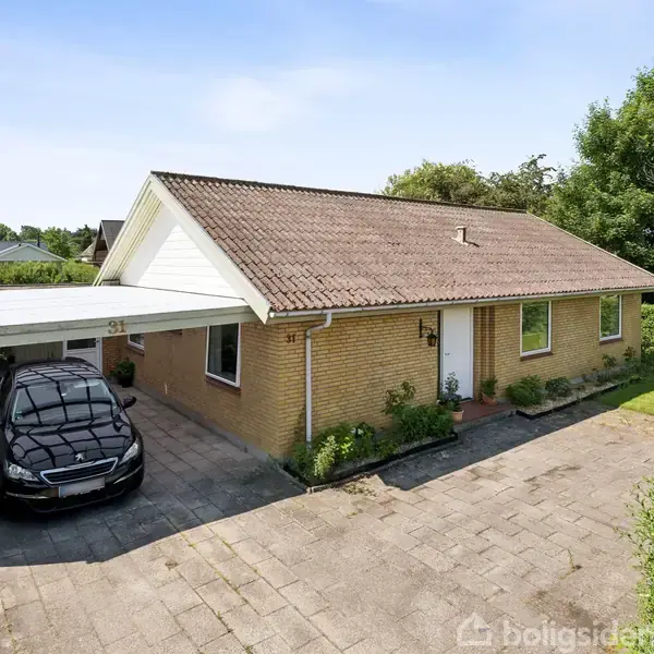 En gul murstensvilla med tegltag står på en flisebelagt indkørsel. En bil er parkeret under en carport. Græsplæne og buske omgiver huset, mens solen skinner. Nummeret "31" er på carporten.