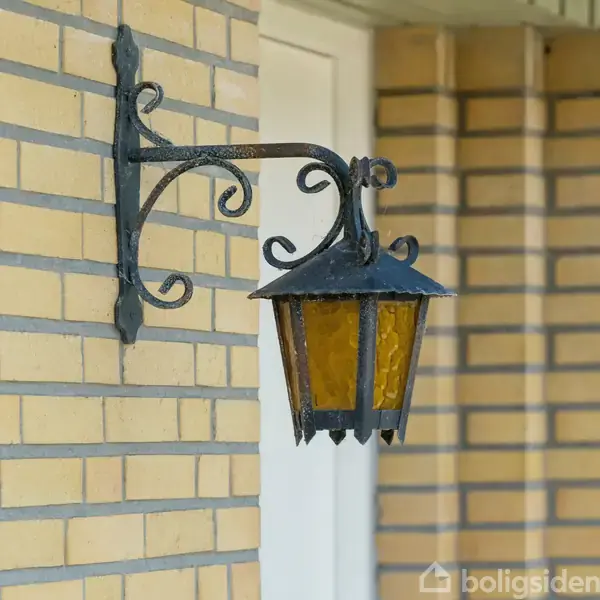 En traditionel udendørs lanterne i sort metal hænger på en murstensvæg. Lampens glas er ravfarvet. I baggrunden ses en dørramme og del af en mur.