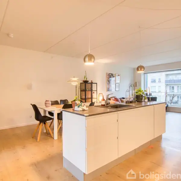 Køkken med hvid køkkenø og hængende lamper, omgivet af spisebord med sorte stole til venstre. Stort vindue i baggrunden giver lys. Boligindretning er moderne og lys.