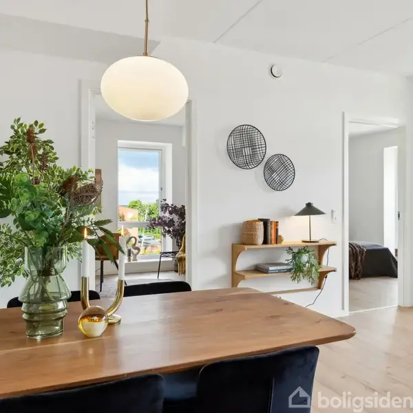 En træbord med en glasvase fyldt med grønne planter står i et lyst rum. En lampeskærm hænger over bordet. I baggrunden ses en hylde med bøger og en lampe.