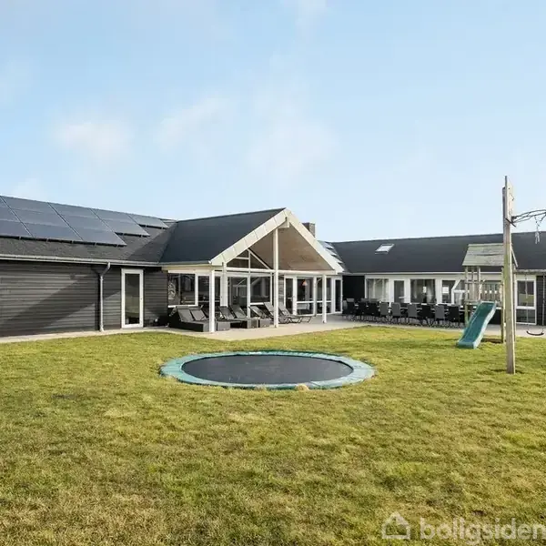 En moderne, sortmalet villa med solpaneler på taget ligger i en grøn have. Foran huset er en nedgravet trampolin og en gynge, der skaber en legepladsstemning.