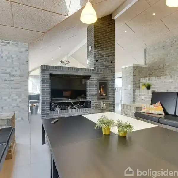 Sort sofabord med små planter står i en moderne stue. Der er grå murstensvægge, to sorte sofaer og en murstenspejs. Rummet er lyst med hængende lamper og store vinduer.