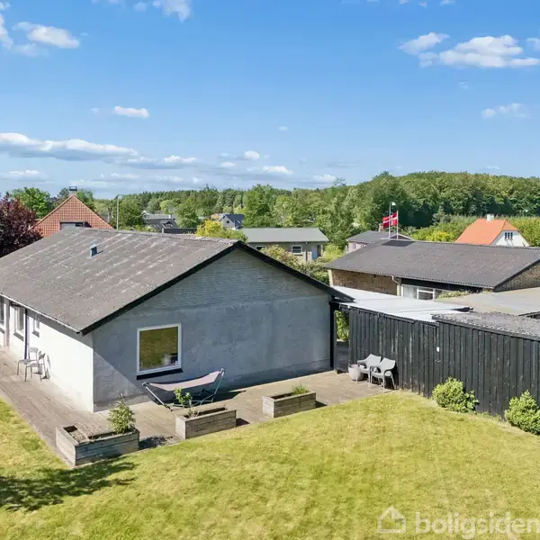 Et gråt hus med fladt tag står i en have. En terrasse har havemøbler og en bænk. Nabolaget er grønt og omgivet af træer under en blå himmel med få skyer.