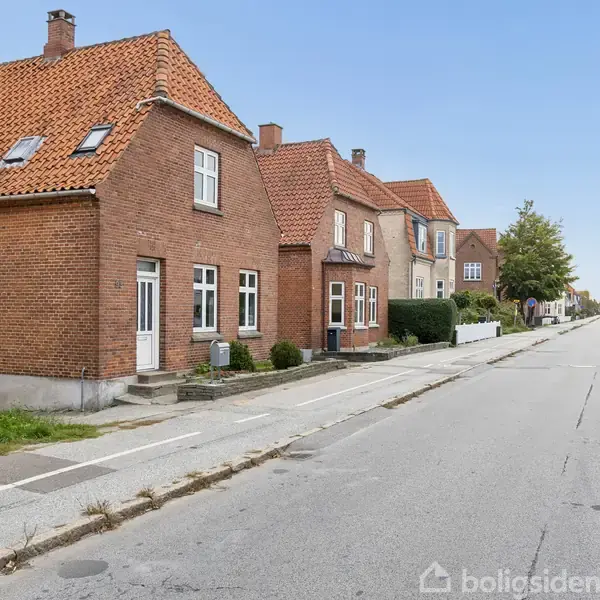 Rødt murstenshus med flere vinduer og skråt tag står ved en stille villavej med parkerede biler i forstadsområde. Træer og flere huse ses længere oppe ad vejen.