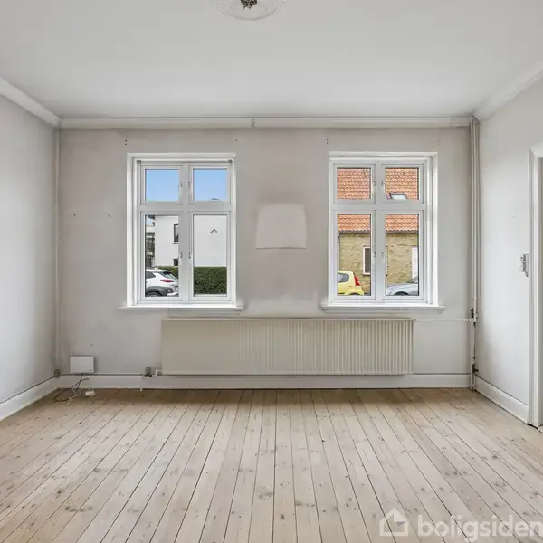 Et tomt rum med trægulv og hvide vægge. To vinduer lukker naturligt lys ind. En radiator er placeret under vinduerne. En dør til højre fører til et andet rum.
