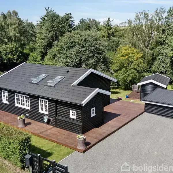 Sort træhus står stille ved siden af en mindre bygning på en stor træterrasse; omgivelserne er naturpræget med mange træer og grus indkørsel.