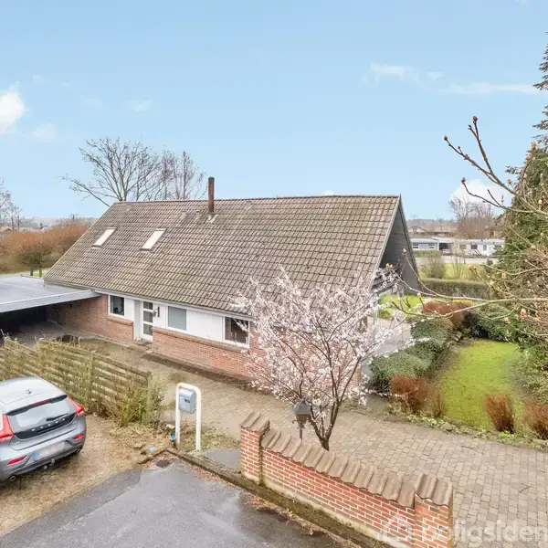 Hus med skråt tag står stille i en have med blomstrende træer, omgivet af en flisebelagt indkørsel. En bil holder parkeret ved siden af en carport.