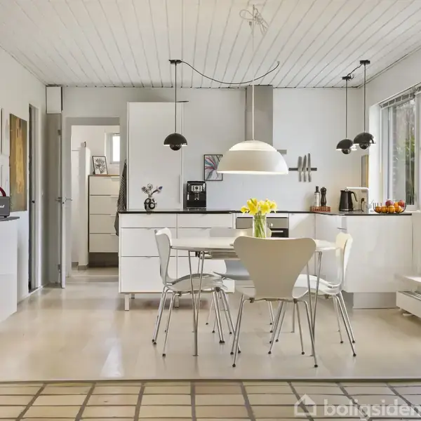 Et lyst, moderne køkken med hvid bordplade. Rundt bord med hvide stole og en vase med blomster. Hængelamper over bordet. Store vinduer til højre med udsigt til haven.