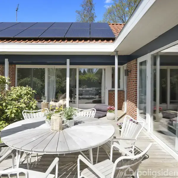 Rundt havebord med stole står på en terrasse omgivet af en murstenshusfacade med store vinduer. Taget har solpaneler, og buske bidrager til en grøn atmosfære.