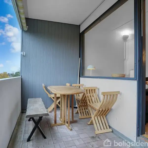 Balkon med rundt træbord og to stole, der står ved siden af en lang bænk. I baggrunden ses en stue gennem et stort vinduesparti på en klar dag.