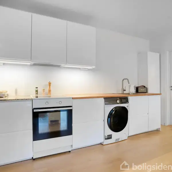 Køkkenet har hvide skabe, en ovn og en vaskemaskine. På bordet står krydderier og en brødrister. Lyset er lyst, og der er et trægulv.