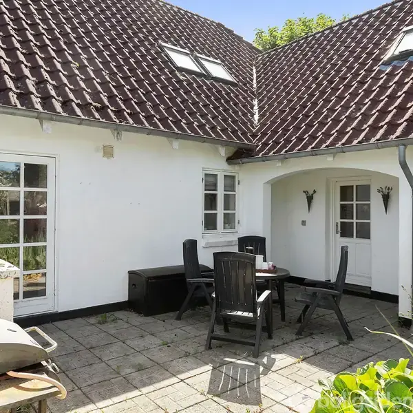 Terrasse med sorte plaststole og bord på flisebelagt område. Omkranset af hvidmalet hus med skråt, mørkerødt tegltag. Flere vinduer og en grill ses i hjørnet. Grønne planter i forgrunden.