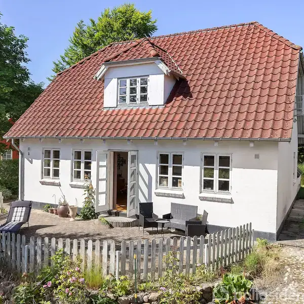 Et hvidt hus med rødt tag står i solen. Huset har tre vinduer og en lille havedør. Foran er en terrasse med møbler, omkranset af et lavt hegn og have.