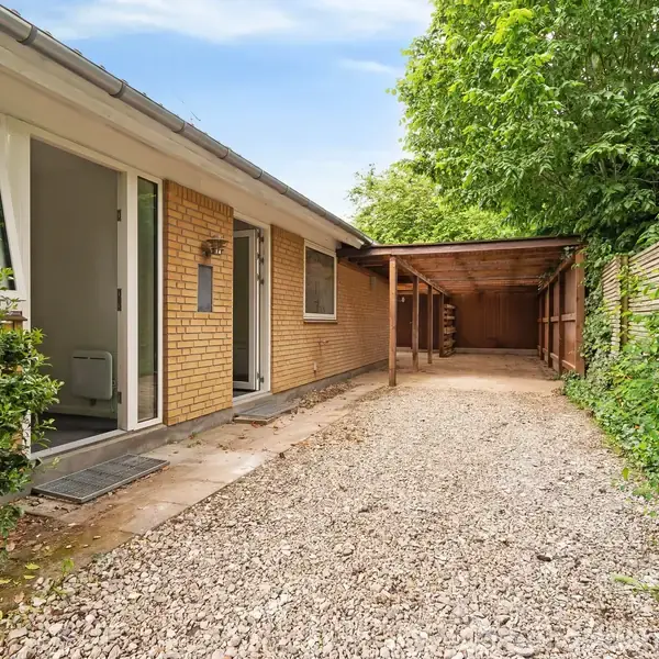 En carport med grusbelagt indkørsel støder op til et gulstenshus med åben dør. Omkringliggende grønne buske og træer skaber en privat atmosfære.