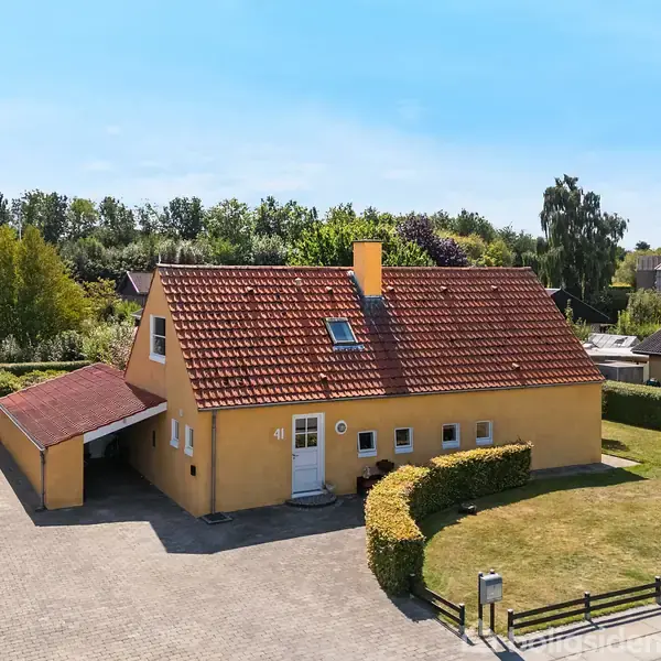 Et gult hus med rødt tegltag står på en flisebelagt plads. En carport sidder til venstre. Baggrund viser grønt hegn og flere huse under blå himmel.