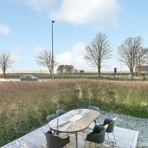 En rund bordgruppe med sorte stole står på en træterrasse. I baggrunden ses en vej, en hæk, og en række nøgne træer under en blå himmel med skyer.