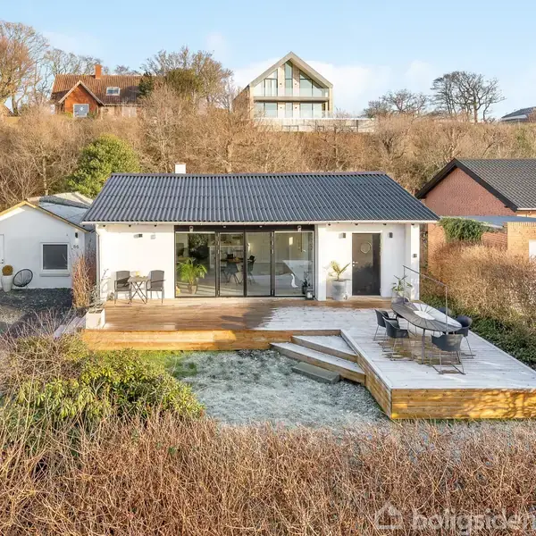 Et moderne hus med store glaspartier og en træterrasse, står stille blandt buske i en beboelsesområde med flere huse i baggrunden.