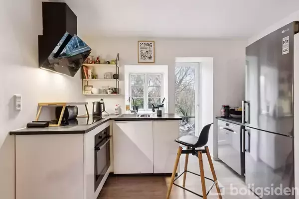 Køkken med hvide skabe og sorte bordplader, indeholder komfur, vask, og køleskab. En barstol står ved en køkkenø. Vindue slipper naturligt lys ind, og nogle hylder bærer køkkenudstyr.