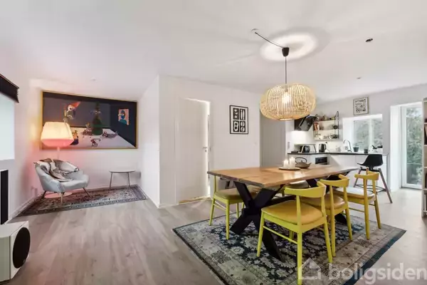 Moderne spisebord med gule stole står under rund pendellampe. Til højre er et køkkenområde med vinduer, og i baggrunden en dekorativ stol og lamper, placeret på et tæppe.