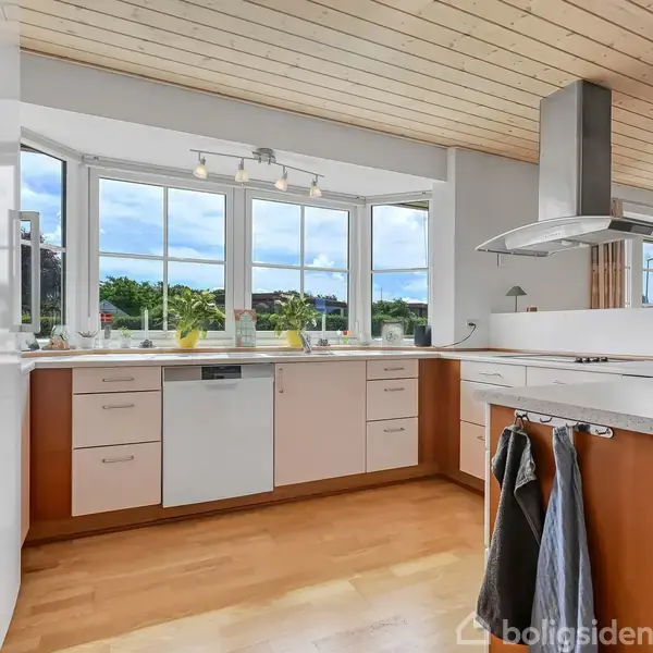 Køkken med træloft og trægulv, hvide skabe og indbygget ovn, omgivet af store vinduer med udsigt over en grøn have. Håndklæder hænger på skabslåger.