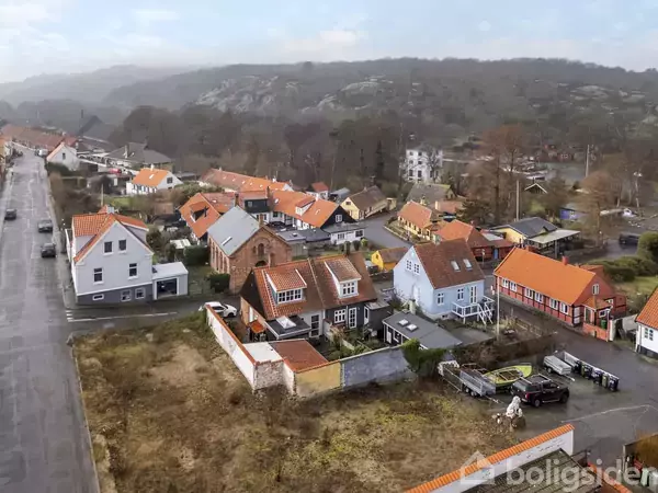 Huse med røde tage ligger tæt sammen. En vej løber langs venstre side med få biler. Omgivelserne rummer marker og bakkede områder i baggrunden.