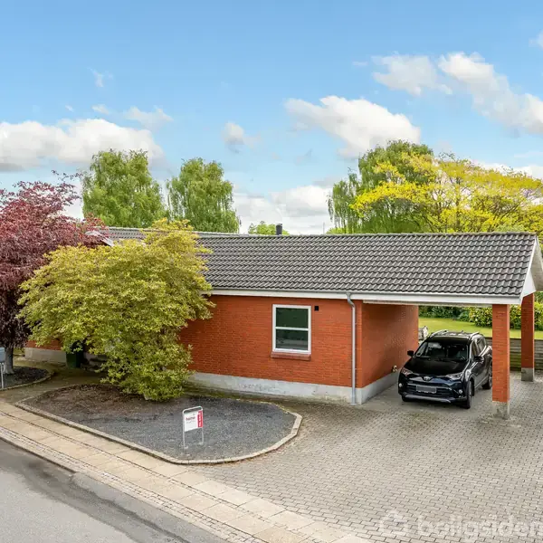Et rødt murstenshus med tilhørende carport, hvor en sort bil holder parkeret. Huset er omgivet af træer og planter, beliggende i en boligkvarter med klar blå himmel.