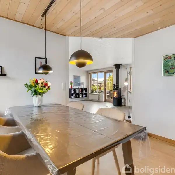 Spisebord med gennemsigtig dug, omkranset af stole, står under to hængelamper. Vase med blomster på bordet. I baggrunden ses stue med brændeovn, bogreol og stort vindue.