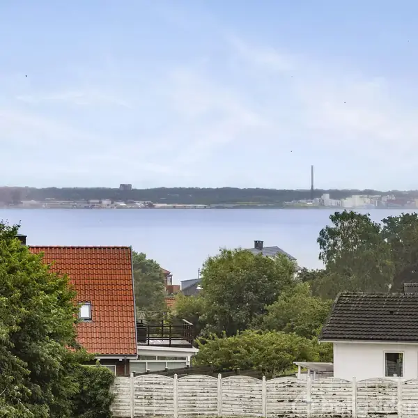 Boliger med røde tage og hvidt stakit ses i forgrunden. Der er en stor vandflade og modsatte kystlinje i baggrunden, omgivet af grønne træer og en blå himmel.