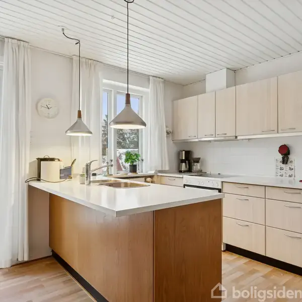 Et køkken med lyse træskabe og en central køkkenø; to pendellamper hænger over. Udenfor ses et snedækket landskab gennem store vinduer og en dør.