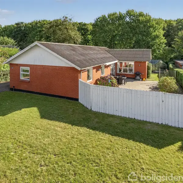 Et rødt murstenshus står stille i en grøn have med en hvid træhegn. Omkring huset er der frodige træer og en terrasse med havemøbler. Tekst: "boligsiden".