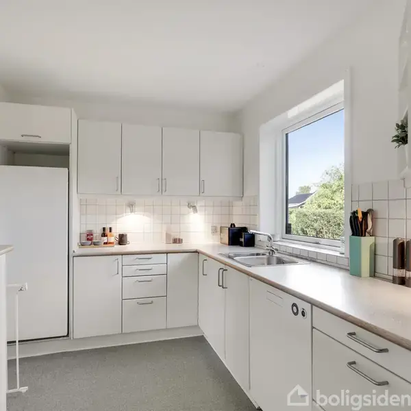 Et funktionelt køkken med hvide skabe og skuffer under en bordplade. Køleskabet står i hjørnet, og der er en stor vindue, der giver naturligt lys. En vask er placeret ved vinduet.