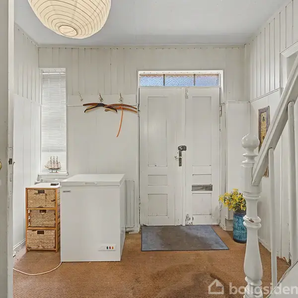 En lys entré med en hvid fryser ved siden af en fletkommode. En væg er dekoreret med en bøjle, mens en vase med blomster står på gulvet ved siden af trappen.