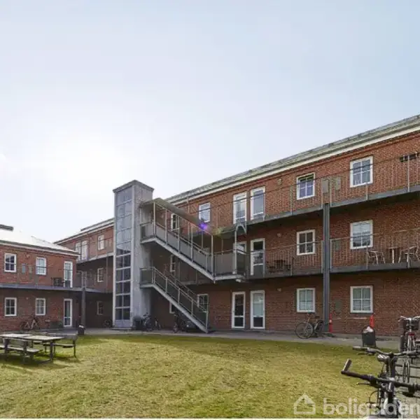 En rød murstensbygning med altaner omkranser en græsklædt gård. Cykler er parkeret langs væggen, og en bænk står midt i gården. Himlen er klar.
