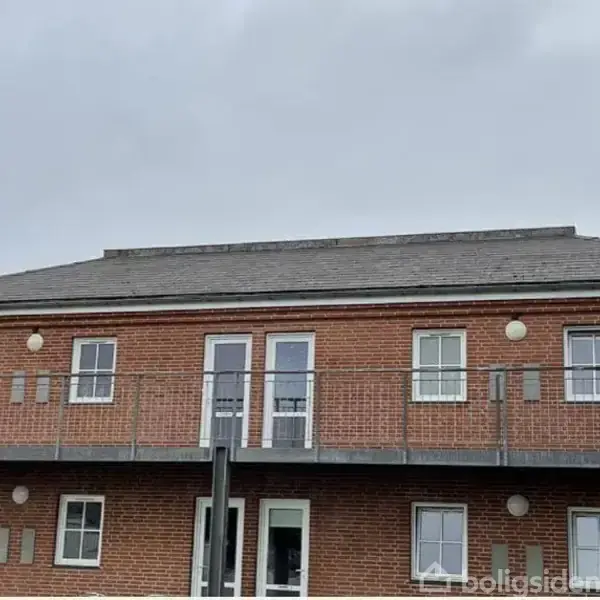Rødt stenbyggeri med to etager, har vinduer og altaner på begge niveauer. Bygningen står under en grå, overskyet himmel. Ingen tekst bortset fra vandmærket.