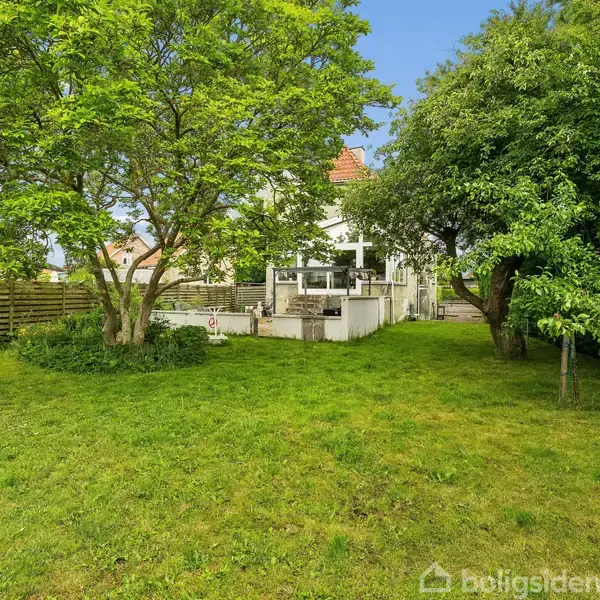 Huset står i baggrunden, omkranset af en frodig have med grøn græsplæne og store træer. En terrasse foran huset skaber et hyggeligt område for udendørs aktiviteter.