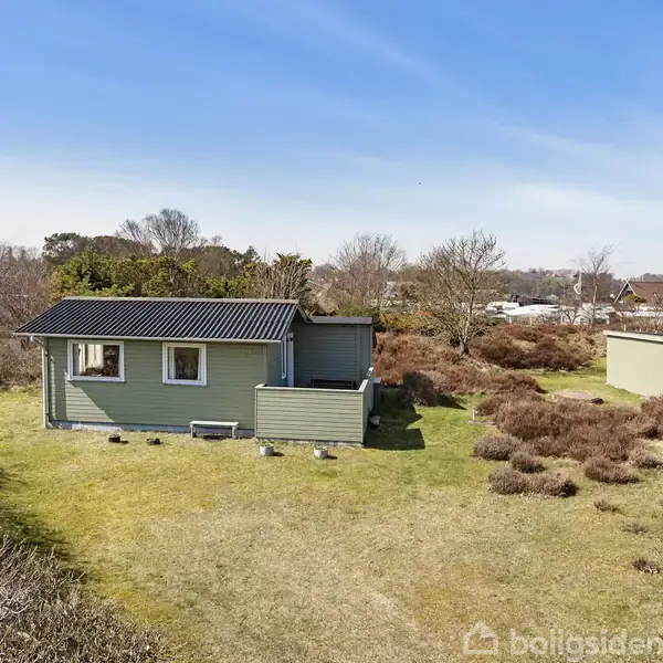 Et lille grønt hus står stille på en åben græsplæne omgivet af lav buskvegetation og løvtræer under en lys blå himmel.