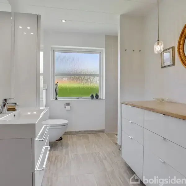 Et lyst badeværelse med hvide skabe, spejl og en vask. Et vindue i baggrunden giver udsigt til natur. Ovalt spejl hænger på højre væg.