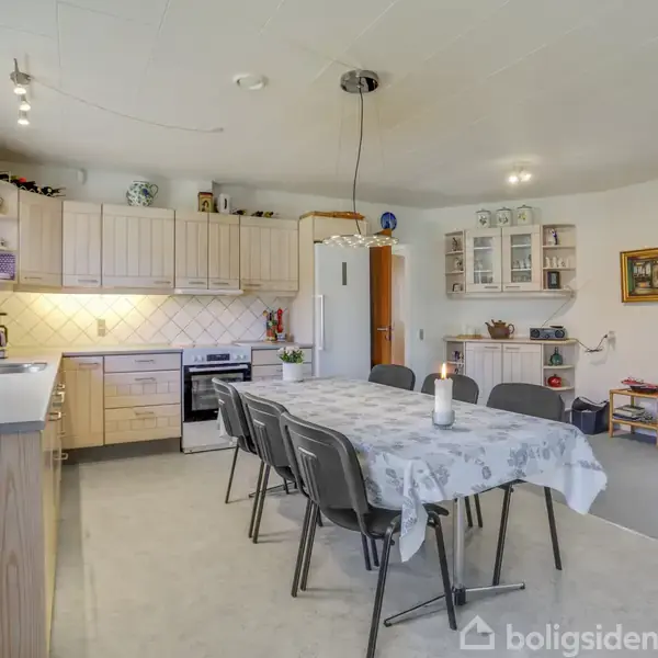 Køkken-alrum med grå spisebordstole og en lys dug, omgivet af træskabe, hvide fliser, og en ovn. Hylder med dekorative genstande og åben dør til tilstødende værelse.