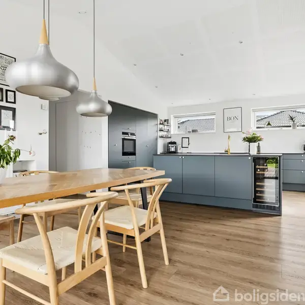 Spisebord med træstole og potteplanter i et moderne køkken. Køkkenø med blå skabe, vinreol og billeder på væggene. Lysekroner hænger fra loftet.