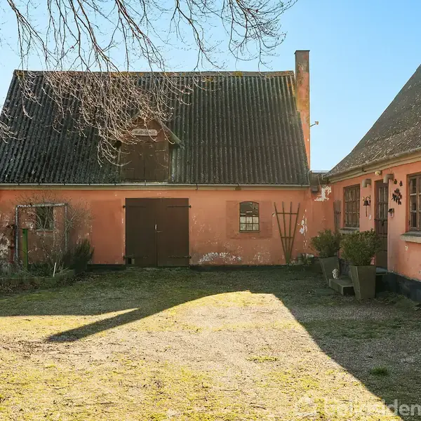 Gammel, lyserød murstensbygning står stille under en klar himmel. Ved siden af ses en mark, og nogle buske vokser tæt på husets vægge i en landlig omgivelser.