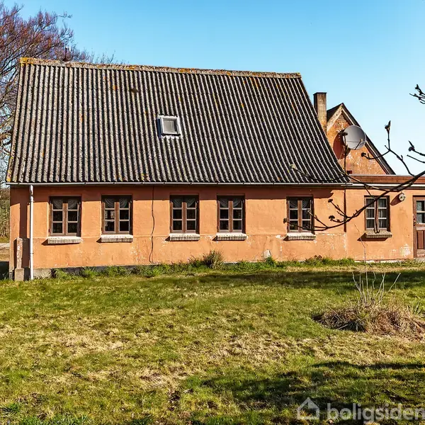 En gammel bygning med skråtag og multiple vinduer står i en grøn have. Træer uden blade ses i baggrunden under en klar, blå himmel.