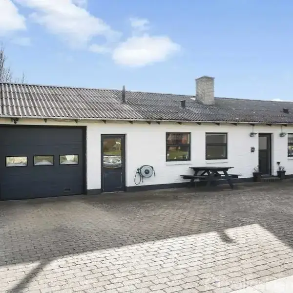 En hvid, étplans villa med sort tag og garage dør, står stille på en brolagt indkørsel. Bord-bænkesæt ses ved siden af huset under en blå himmel.