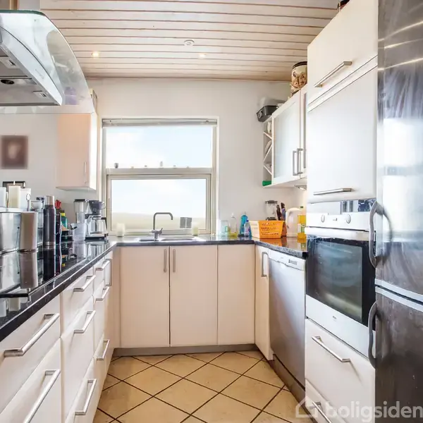 Et moderne køkken med hvide skabe og sorte bordplader. Et stort køleskab står til højre. Et vindue over vasken giver naturligt lys i rummet.