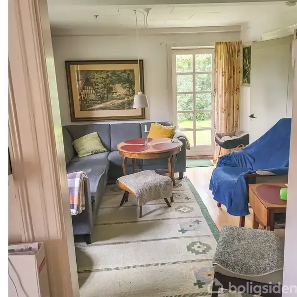 Stue med to sofaer og et bord, dekoreret med puder og tæppe. Et maleri hænger på væggen. Store vinduer giver udsigt til have. En blå stofdækning findes på en stol.