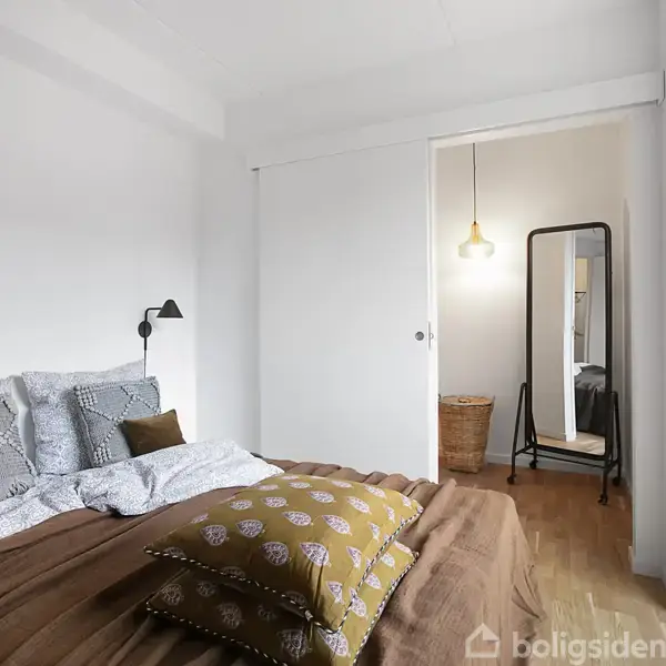 Soveværelse med en seng til venstre dækket med brune og mønstrede puder, en væglampe ovenfor, og et gulvspejl i hjørnet ved en hængende lampe i lys omgivelser.