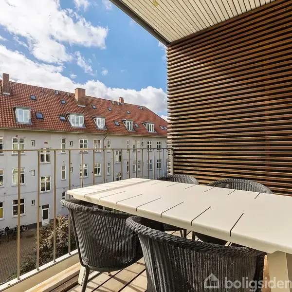 Bord og stole står på en balkon, omgivet af en træbeklædning. Udsigt over hustage med røde tegl og himmel med spredte skyer ses i baggrunden.